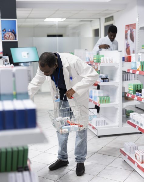 African American pharmacist organizing pharmacy shelves with medication, checking packaging and controlling stock levels to ensure order, cleanliness and efficiency in the pharmacy interior.